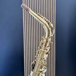 Close-up of a gold saxophone neck with cork pad and keys against a blue striped background, highlighting the instrument detail.