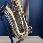 Close-up of a polished brass saxophone with ornate engravings resting in a blue stand on a light wooden surface against a blue wall.