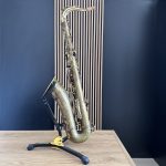 Bronze-tone saxophone resting on a black-and-yellow stand on a light wood table, against a blue wall with vertical wooden slats behind it.