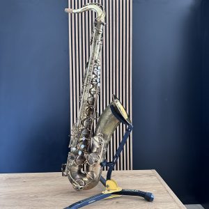 Brass saxophone resting diagonally on a wooden table with a blue wall behind and a yellow Hercules stand visible at the base.