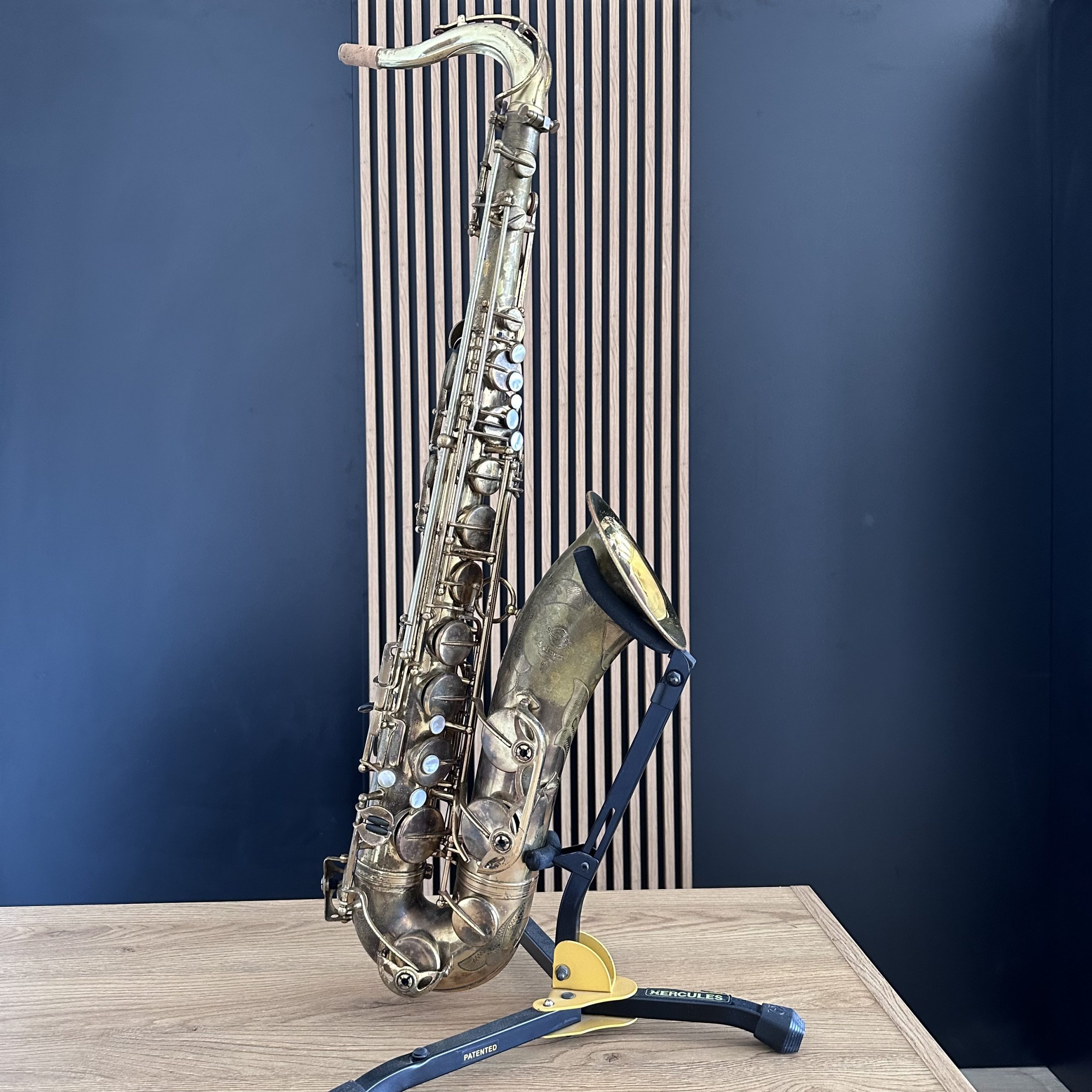 IMG_0158 Brass saxophone resting diagonally on a wooden table with a blue wall behind and a yellow Hercules stand visible at the base.