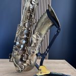Close-up of a tarnished silver saxophone resting on a yellow stand on a light wooden floor, with a dark blue wall behind it.