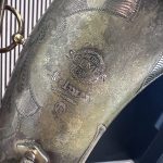 Close-up of a weathered metal medallion with laurel engraving and rope-edge border, partially showing text.