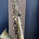 Close-up of a weathered brass saxophone neck with keys and rods against a dark blue background.