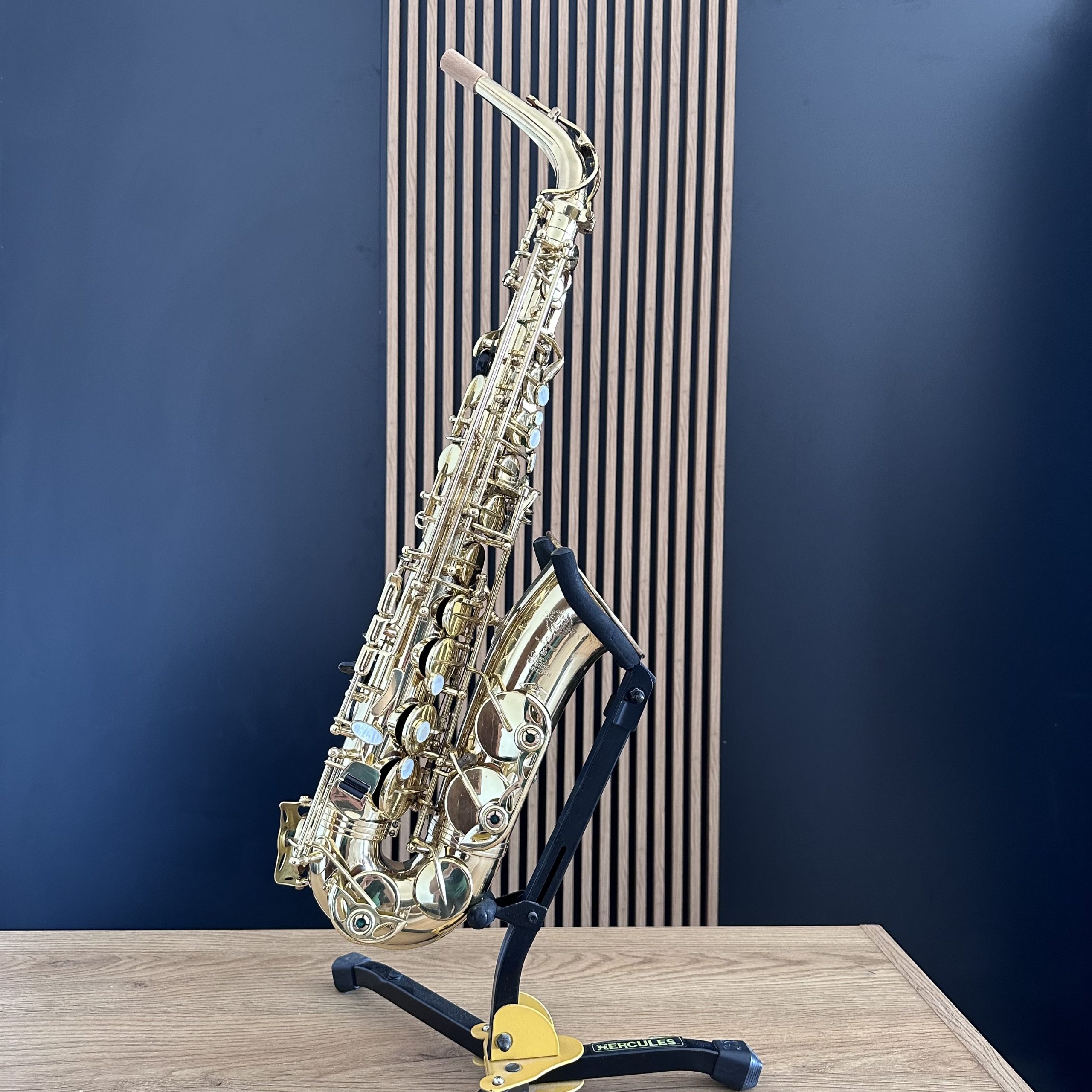 IMG_9611 Gold saxophone resting in a black-yellow stand on a light wooden table against a dark blue wall with vertical slats behind it.