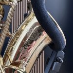Close-up of a polished gold saxophone showing engraved crest and a dark velvet strap nearby on a blue background.
