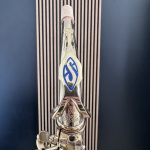 Close-up of a polished metal lever or tool with a blue diamond emblem, set against a dark striped background.