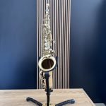 Brass saxophone resting on a black stand on a wooden table with a blue studio backdrop behind it.