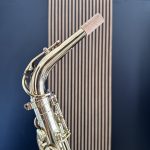 Close-up of a shiny saxophone neck and mouthpiece with a blue background and striped wall behind it, highlighting metal details and cork tip.