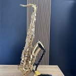 Gold alto saxophone resting on a stand in a recording studio, against a blue wall with vertical wooden panels and a light wooden table nearby