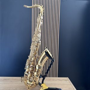 Gold alto saxophone resting on a stand in a recording studio, against a blue wall with vertical wooden panels and a light wooden table nearby