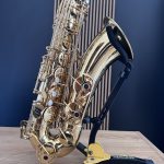 Close-up of a gold saxophone on a black stand, showing intricate keys and polished brass surface against a blue wall and wooden floor background.