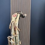 Close-up of a shiny chrome sculpture of a standing human figure, reflecting light, against a blue wall with vertical slats behind it.