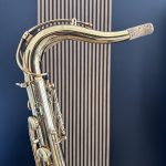 Close-up of a shiny brass saxophone neck with cork mouthpiece against a dark blue background.