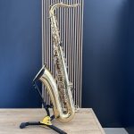 Golden brass saxophone resting on a yellow stand on a light wooden table, against a blue wall with vertical panels.