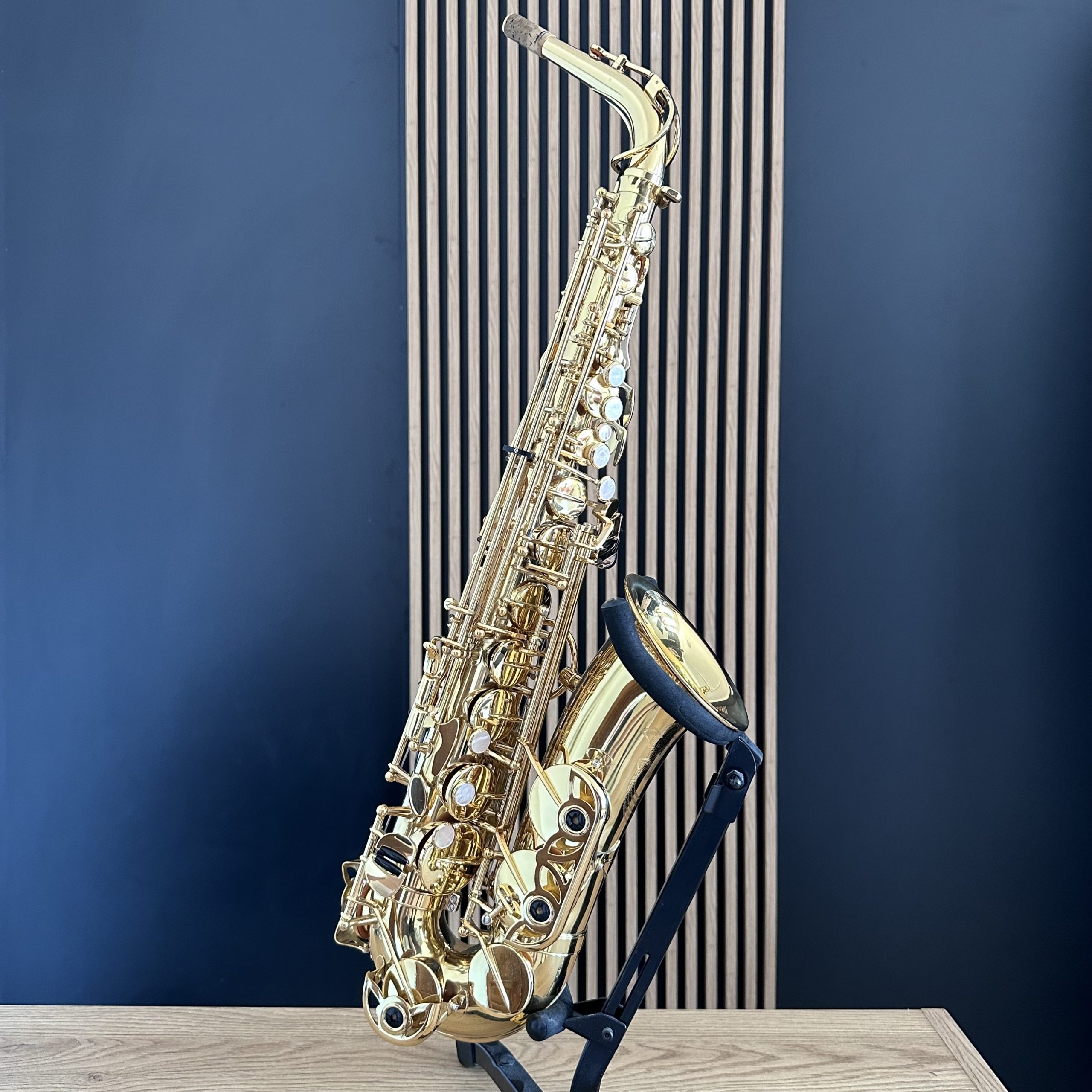 IMG_0197 Shiny gold saxophone resting on a black stand against a blue wall and light wood floor in a studio setting.