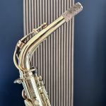 Close-up of a gleaming brass saxophone neck with cork pad and tuning mechanism, against a blue striped background.
